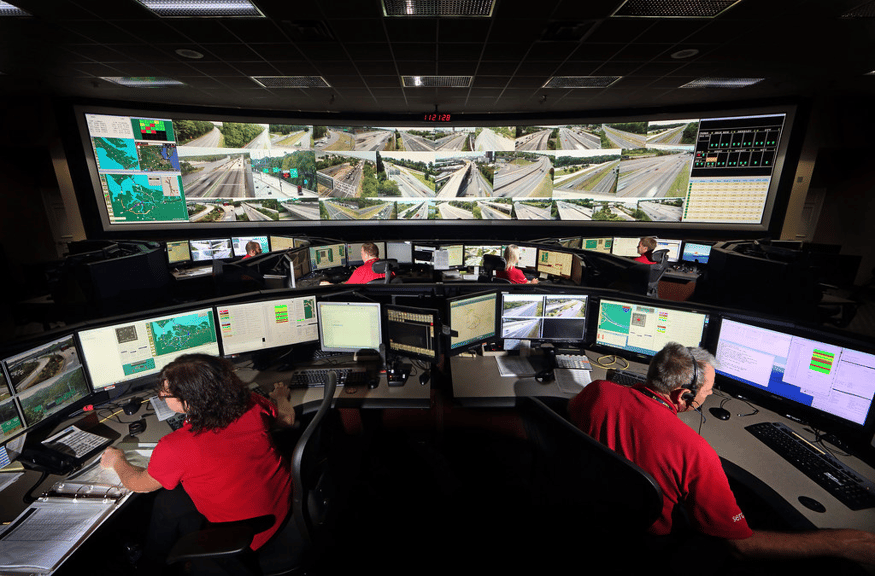 一对男女在安全操作室的工作站上工作，他们身后的视频墙显示着交通摄像头的实时画面。