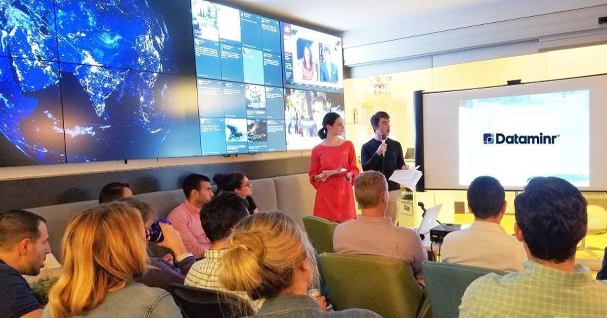 Dataminr employees in a meeting room sitting opposite a projector, and beside a video wall with social media and news updates