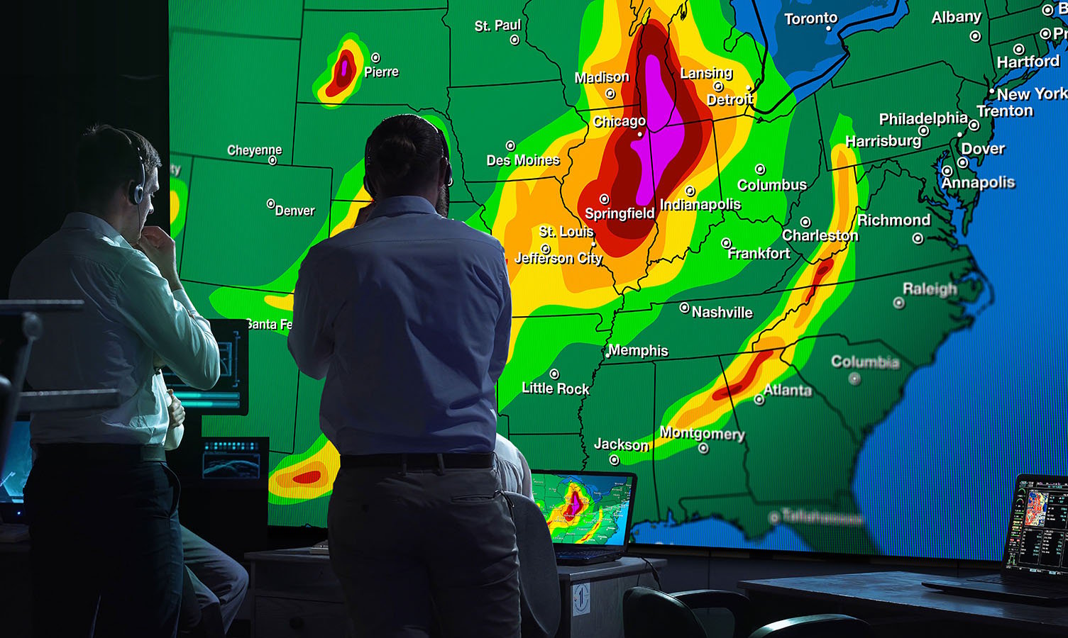 2名男子在天气监测控制室，看着显示美国天气图的大型视频墙。