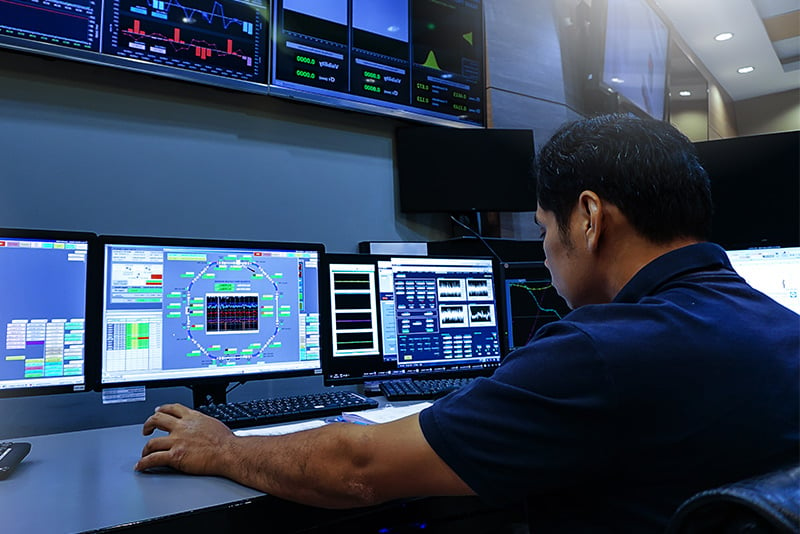Network operations center employee looking down at notes on his desk while sitting, with video walls displaying data dashboards